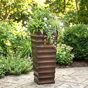 May include: A tall, bronze-colored, rectangular planter with a textured, ridged design. The planter is filled with a variety of green plants, including ferns, ivy, and flowering plants. The planter is outdoors, surrounded by greenery.