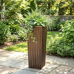 May include: A tall, rectangular, brown wooden planter filled with various green plants and foliage. The planter has vertical ribbed detailing and sits on a stone patio. The background features lush greenery and a stone wall.