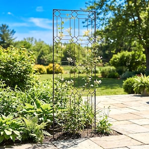 May include: A black metal garden trellis with a geometric design, standing upright in a garden setting. The trellis is adorned with climbing plants with white flowers, set against a backdrop of lush greenery and a blue sky.