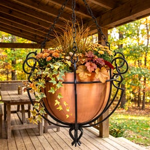 May include: A copper-colored bowl planter held by a black metal frame with scrollwork details and a hanging chain. The planter contains a variety of plants including orange flowers, green and yellow ivy, and brown foliage, creating a full arrangement.