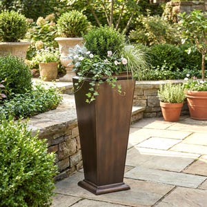 May include: A tall, bronze-colored outdoor planter filled with greenery and flowers. The planter has a square base and a tapered, rectangular body. The plants include small white flowers, trailing vines, and ornamental grasses. The background features other planters and lush landscaping.