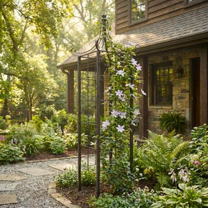May include: A tall, dark metal garden trellis with a decorative top, supporting climbing plants with light purple flowers. The trellis is set in a lush garden with a stone pathway and a house in the background.