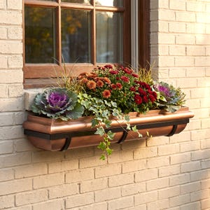 May include: A rectangular copper window box planter mounted on a light-colored brick wall. The planter is filled with a vibrant arrangement of deep red and orange chrysanthemums, purple ornamental kale, and trailing green ivy foliage.