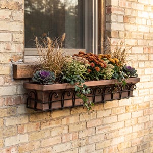 May include: A rectangular copper-toned window box planter held by a black wrought iron frame with a decorative scroll pattern. The planter is filled with a variety of plants, including ornamental kale, tall grasses, ivy, and orange-toned flowers.