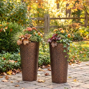 May include: Two tall, cylindrical planters with a hammered copper-toned finish. Each planter is filled with a vibrant arrangement of orange and yellow flowers, green foliage, and trailing ivy, creating a lush display of autumn-inspired colors.