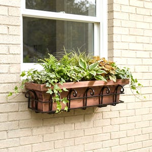 May include: A rectangular copper-colored window box planter encased in a black metal frame with decorative scrollwork. The planter is filled with a variety of green foliage, including ferns, hostas, and trailing ivy, mounted on a brick wall.