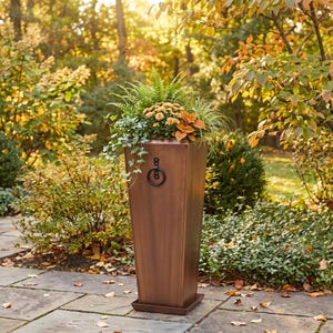 May include: A tall, tapered, copper-colored metal planter with a square base and a matching drip tray. The planter features a black metal ring handle attached to the front. It is filled with a variety of green ferns, ivy, and orange flowers.