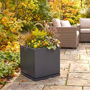 May include: A square, dark gray planter box containing a variety of plants, including yellow flowers, green foliage, and tall ornamental grasses. The planter sits on a flat base and is positioned on a stone patio surface outdoors.