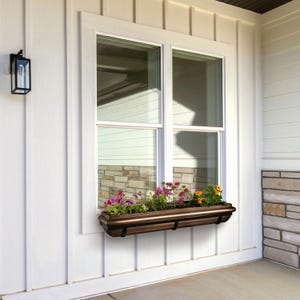 May include: A brown window box planter with a decorative trim, filled with colorful flowers, is mounted below a white window on a white siding exterior.
