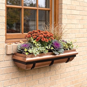 May include: A rectangular copper window box planter with black metal support bands, filled with orange chrysanthemums, purple ornamental kale, green and white variegated ivy, and tall ornamental grasses, mounted on a tan brick wall.