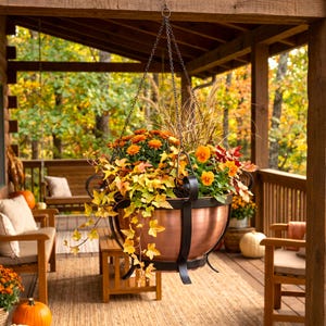 May include: A copper-colored metal hanging planter bowl with a black iron frame and chain, filled with a vibrant autumn arrangement of orange chrysanthemums, yellow ivy, tall ornamental grasses, and orange pansies in a rounded basin.