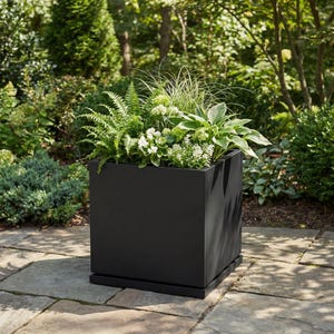 May include: A black square planter box filled with a variety of green plants and white flowers. The planter sits on a matching black base, and is placed on a stone patio. The background features lush green foliage.