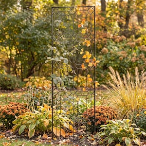 May include: A tall, rectangular black metal garden trellis featuring a geometric grid pattern. A climbing vine with green leaves and some yellowing autumn foliage is growing up the vertical structure, which stands upright in a garden setting.