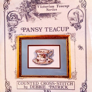 May include: A framed cross-stitch of a teacup and saucer with floral details. The artwork is labeled "Pansy Teacup" from the "Victorian Teacup Series." The frame is gold-toned, set against a light blue background, with decorative borders.