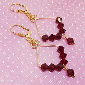 May include: A pair of gold earrings with a geometric design. Each earring features a triangle shape with a dangling row of red crystal beads.
