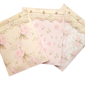 May include: Three decorative envelopes with a floral pattern of pink roses and green leaves. Each envelope has a lace trim and pearl accents.