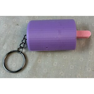 May include: A purple popsicle-shaped keychain with a pink stick. The keychain is attached to a silver key ring and chain. The popsicle is a solid color and has a smooth texture.