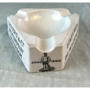 May include: White ceramic ashtray with a triangular shape and three resting areas. The ashtray features text that reads "LEVERT & CO DE WILDEMAN AMSTERDAM" and "LIKEUREN DE WILDEMAN". It also has a graphic of a man and the year "1690".