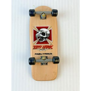 Pode incluir: Um skate em miniatura com um deck castanho claro, rodas pretas e ferragens prateadas. O deck apresenta um gráfico vermelho e branco de uma caveira com o texto "TONY HAWK" e "POWELL PERALTA".