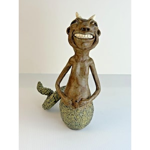May include: A brown ceramic sculpture of a creature with a wide, toothy grin and two small horns on its head. The upper body is smooth and brown, while the lower body and tail feature a textured, cross-hatched pattern in a muted green tone.