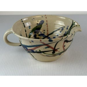 May include: A ceramic bowl with a handle and a spout, featuring a beige base color. The bowl is decorated with colorful abstract paint splatters in blue, green, red, and black. The bowl has a rounded shape and a smooth finish.