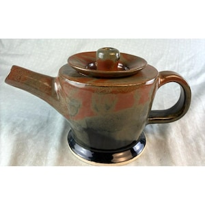 May include: A handcrafted ceramic teapot with a dark brown, red, and black glaze. The teapot features a spout, handle, and a lid with a small, cylindrical knob. The base is black and the teapot is designed for brewing tea.
