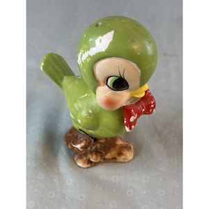 May include: A vintage ceramic bird figurine, primarily green with white accents, perched on a brown base. The bird has a round head, a red bow tie with white polka dots, and a yellow beak. The bird's eyes are black with green and white details.