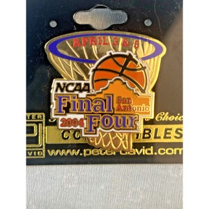 May include: A gold-colored pin featuring a basketball and hoop design, with the text "NCAA Final Four 2004 San Antonio." The pin also includes the dates "April 3 & 5." The pin is attached to a black backing.