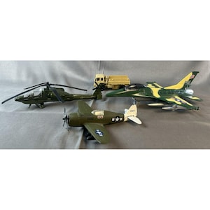 May include: A collection of toy military vehicles, including a green helicopter, a tan truck, a green and yellow fighter jet, and a green propeller plane. The toys are arranged on a gray surface.
