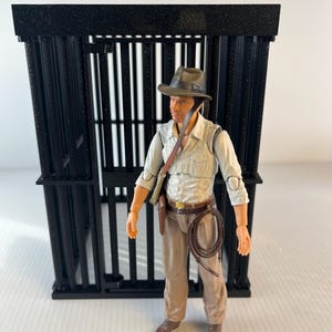 May include: A black plastic cage with a figure wearing a brown hat, a light brown shirt, a brown belt, and tan pants. The figure is holding a whip in their right hand.