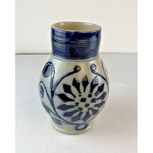 May include: A stoneware vase with a light gray body and a dark blue glazed neck featuring horizontal ridges. The exterior is decorated with a dark blue hand-painted floral motif consisting of a central sunflower-like bloom and swirling vines.