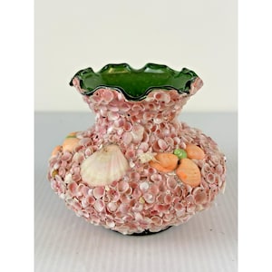 May include: A decorative vase covered in small pink seashells. The vase has a ruffled green rim and is adorned with larger seashells and orange accents. The base is black.