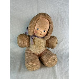May include: Vintage stuffed doll with a painted face, blue eyes, and a brown fur hood. The doll has a light brown body and limbs, with a lavender ribbon tied around its neck. A classic collectible toy.