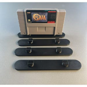 Pode incluir: Um suporte de plástico preto com três níveis para exibir cartuchos de jogos Super Nintendo. O nível superior tem um cartucho de jogo Super Nintendo cinza com o título "Illusion of Gaia" no rótulo.