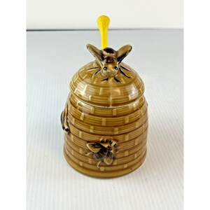 May include: A ceramic honey pot shaped like a beehive with a brown, textured, woven-look finish. The lid features a sculpted bee and a bright yellow handle. A small bee is also sculpted onto the side of the pot, which has a glossy glaze.