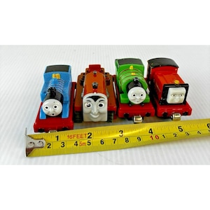 May include: Four toy train engines in blue, brown, green, and red, each with a smiling face. The trains are on red bases. A yellow measuring tape shows the length of the trains.
