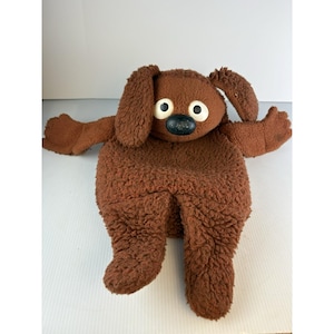May include: A brown plush dog toy with a textured, fuzzy fabric surface. The toy features two large, round white eyes with black pupils and a prominent, rounded black nose. It has floppy ears, outstretched arms, and two legs extending downward.