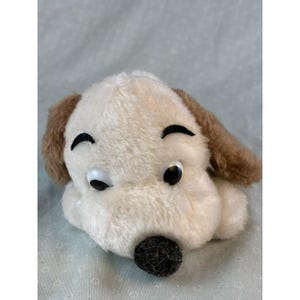 May include: A plush dog toy with a cream-colored body, brown ears, and a black nose. The toy has large, round eyes and curved black eyebrows, giving it a friendly appearance. The toy is lying on a light blue surface.