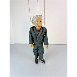 May include: A vintage string puppet of Albert Einstein, dressed in a dark suit with a black trim. The puppet has a painted face with a mustache and white hair. The puppet is suspended by strings.