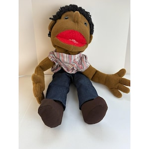 May include: A plush hand puppet featuring a brown fabric body, black yarn hair, and a bright red mouth. The puppet is dressed in a striped shirt with thin multicolored lines and dark blue denim-style pants, with brown fabric feet attached.