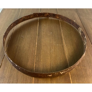 May include: A rusty, circular metal band, likely from a barrel or similar container, sits on a wooden surface. The band is a dark brown color, showing signs of age and wear. The wood grain is visible.