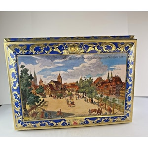 May include: A rectangular metal tin featuring a vibrant historical illustration of a Nuremberg town square with timber-framed buildings, trees, and figures on horseback. The border is decorated with ornate gold filigree on a deep blue background.