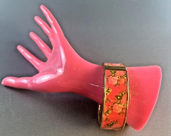 May include: A gold bangle bracelet with a pink floral pattern. The bracelet is on a pink mannequin hand.