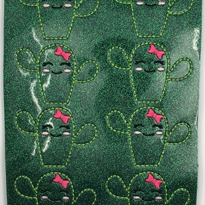 May include: A dark green glitter sheet featuring eight embroidered cactus motifs. Each cactus has a smiling face, pink cheeks, and a small pink bow. The cacti are outlined with light green stitching and arranged in two vertical columns of four.