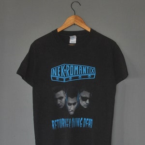 May include: Black t-shirt featuring a blue graphic of the band Nekromantix. The graphic shows the band's name in a coffin-shaped box with skulls and the text "Return of the Living Dead".