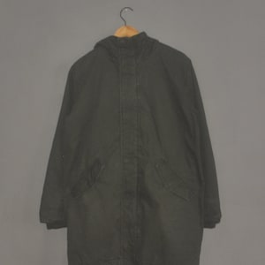 Peut inclure: Parka vert foncé avec capuche, deux poches avant et cordon de serrage à l'ourlet. L'intérieur est beige clair. La veste est suspendue à un cintre en bois sur fond gris.