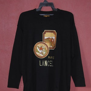 May include: Black long-sleeve shirt with a gold embroidered design featuring two images of buildings and the text "Paris Lancel".