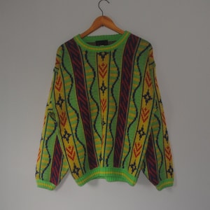 May include: A colorful sweater with a geometric pattern. The sweater features vertical stripes in green, yellow, and red, with a ribbed collar and cuffs. The sweater is hanging on a wooden hanger.
