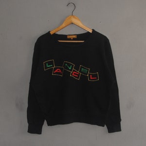 May include: Black crew neck sweatshirt with the word "LANEL" embroidered in a colorful, geometric design. The letters are outlined in gold, with green, red, and yellow accents. The sweatshirt is hanging on a wooden hanger against a gray background.