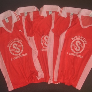 Op de afbeelding: Vijf rood-roze voetbalshirts met witte letters. De shirts hebben een witte kraag en witte strepen aan de zijkanten. De tekst op de shirts luidt "Wasser Heizung H. Schneitzlinger".